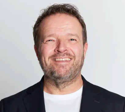 Porträtt av en smilende mann i jakke og t-skjorte, med kort hår og skjegg.